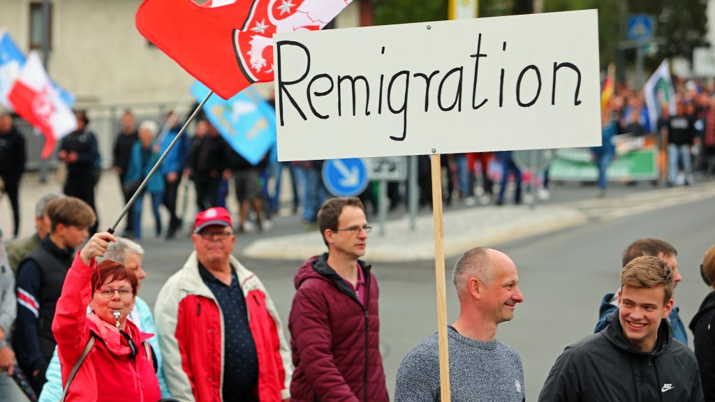 Mehrere Menschen bei einer extrem rechten Demonstration in Schleusingen. eine Fraus schwenkt eine Thüringen-Fahne. Neben ihr läuft ein Mann mit einem Schild und der Aufschrift Remigration
